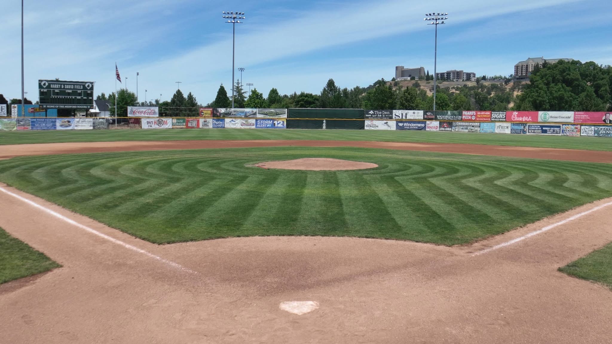 View from behind home plate at Harry & David Field toward the outfield scoreboard and sponsor banners in Medford, Oregon.